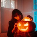 A woman carves a glowing jack-o'-lantern indoors amidst cobwebs and eerie lighting, celebrating Halloween.